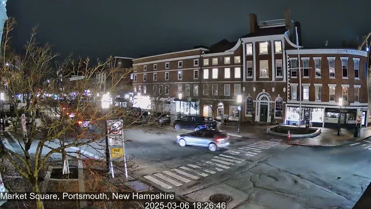 A nighttime street scene shows several cars at an intersection in front of brick buildings with illuminated windows.