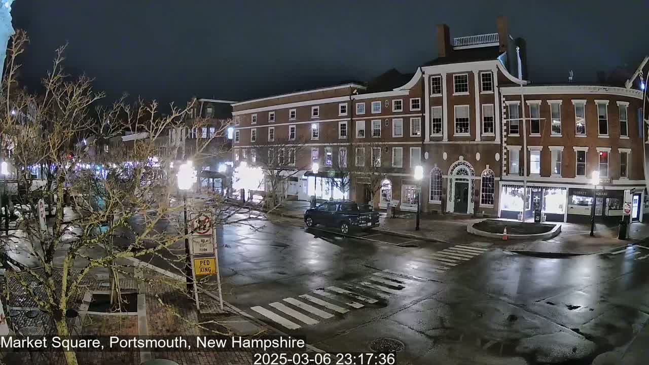 A nighttime view of a mostly empty town square with wet streets and bare trees under a dark sky.
