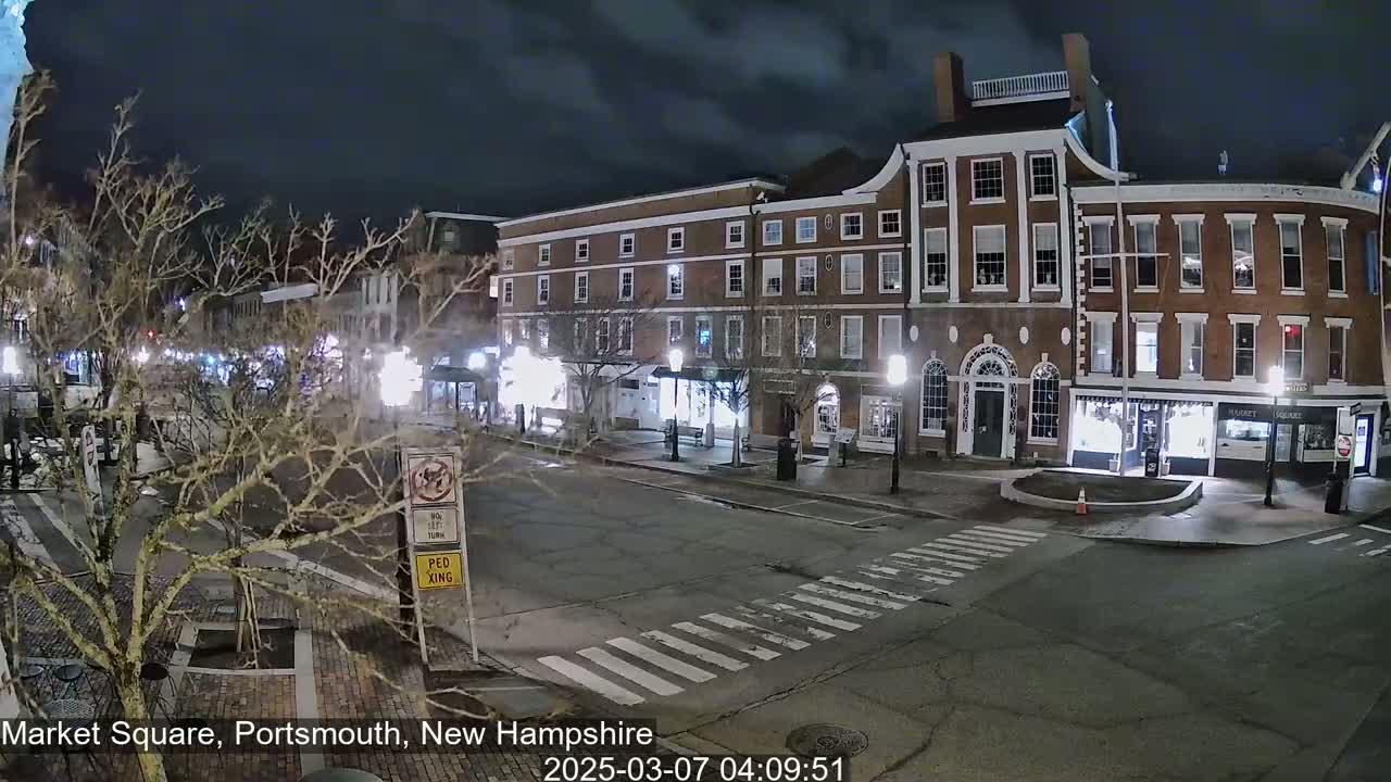 A nighttime view of a mostly empty town square with brick buildings, streetlights, and bare trees under a dark sky.