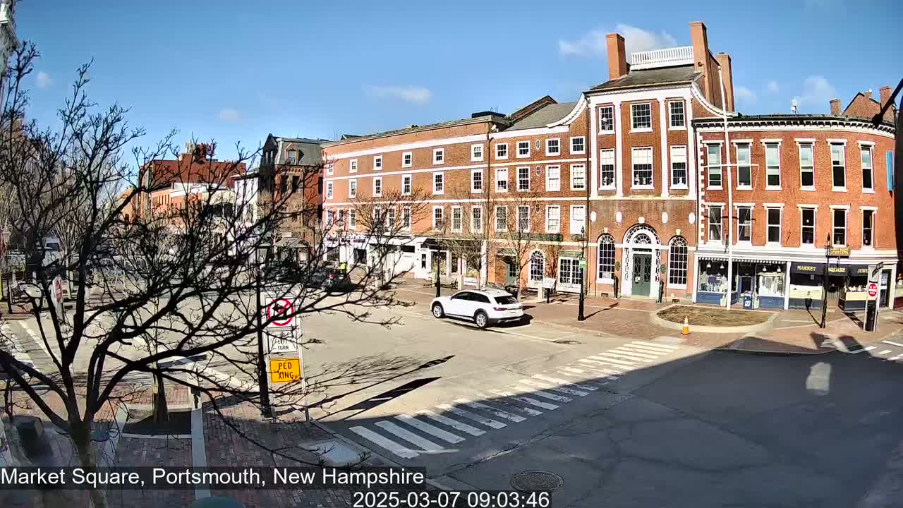 A sunny day in a brick-lined city square with a few cars and bare trees.