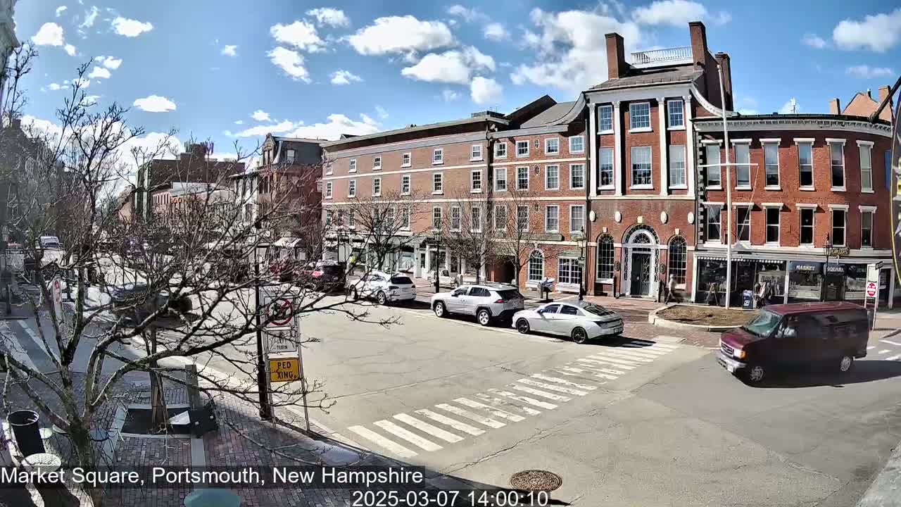 A sunny day in a city square shows several brick buildings, some bare trees, and several cars parked and driving around a crosswalk.