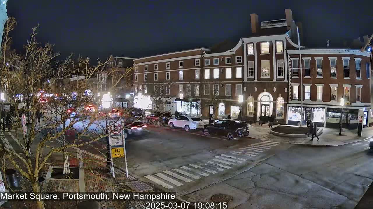 A nighttime street scene shows several cars driving through a town square in front of brick buildings with illuminated windows under a clear, dark sky.