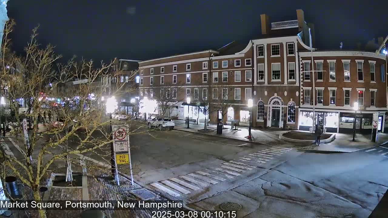 A nighttime view of a mostly empty town square with brick buildings, sparse traffic, and bare trees under a dark sky.