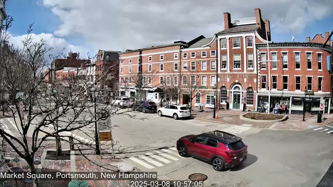 A partly cloudy day reveals a red car driving through a town square lined with brick buildings and bare trees.