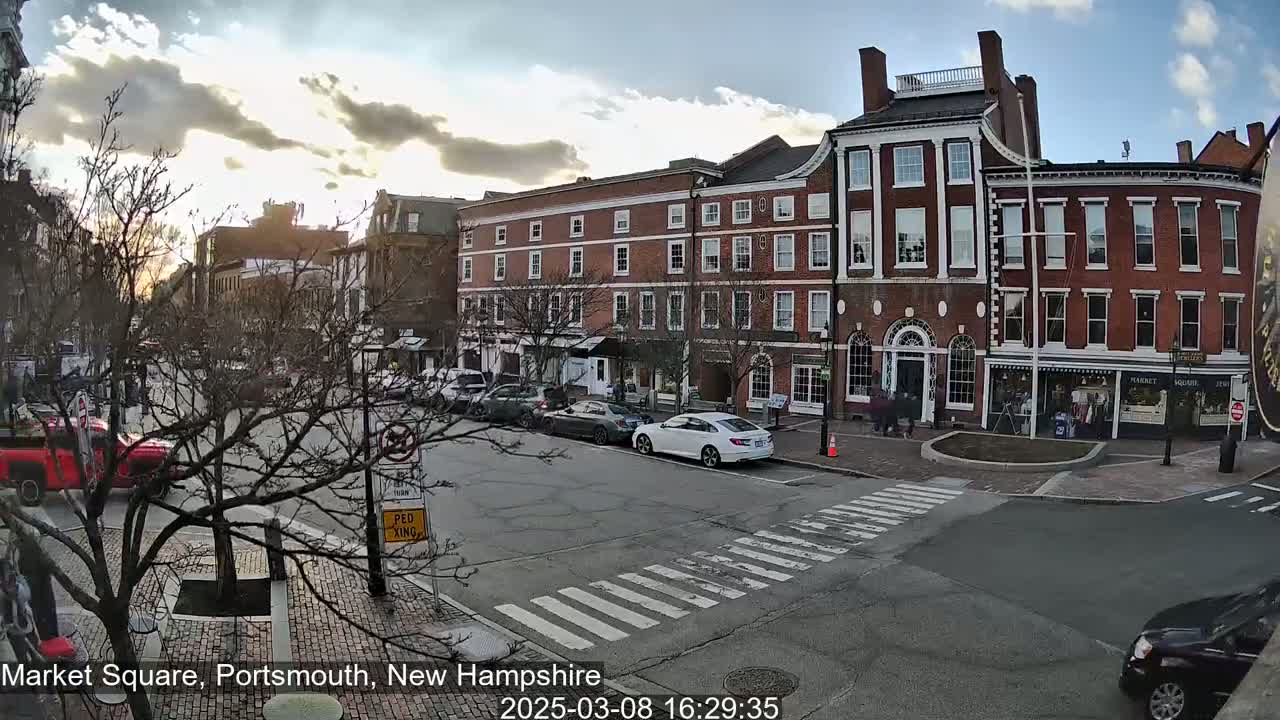 A mostly sunny street scene shows several brick buildings, parked cars, and a crosswalk in a town square.