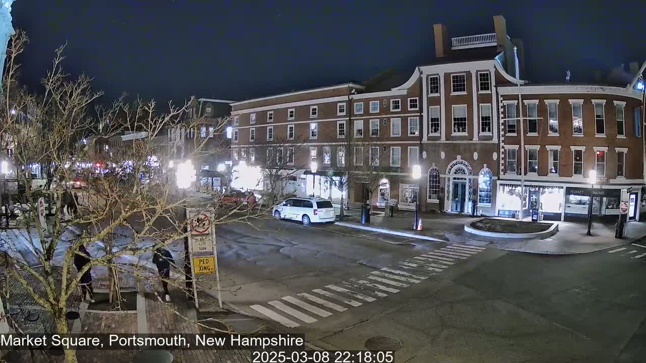 A nighttime view of a mostly empty town square with brick buildings, streetlights, and a few pedestrians and vehicles.