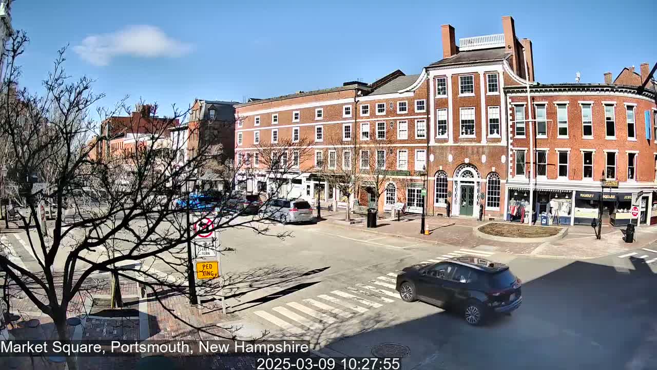 A sunny day reveals a brick-lined town square with several parked cars and one driving across a crosswalk, surrounded by multi-story buildings.
