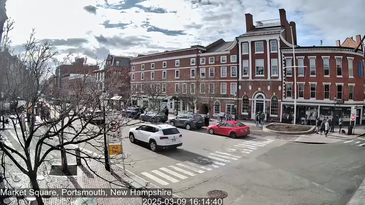 Under a partly cloudy sky, cars drive through a city square lined with brick buildings, and pedestrians walk on the sidewalks.