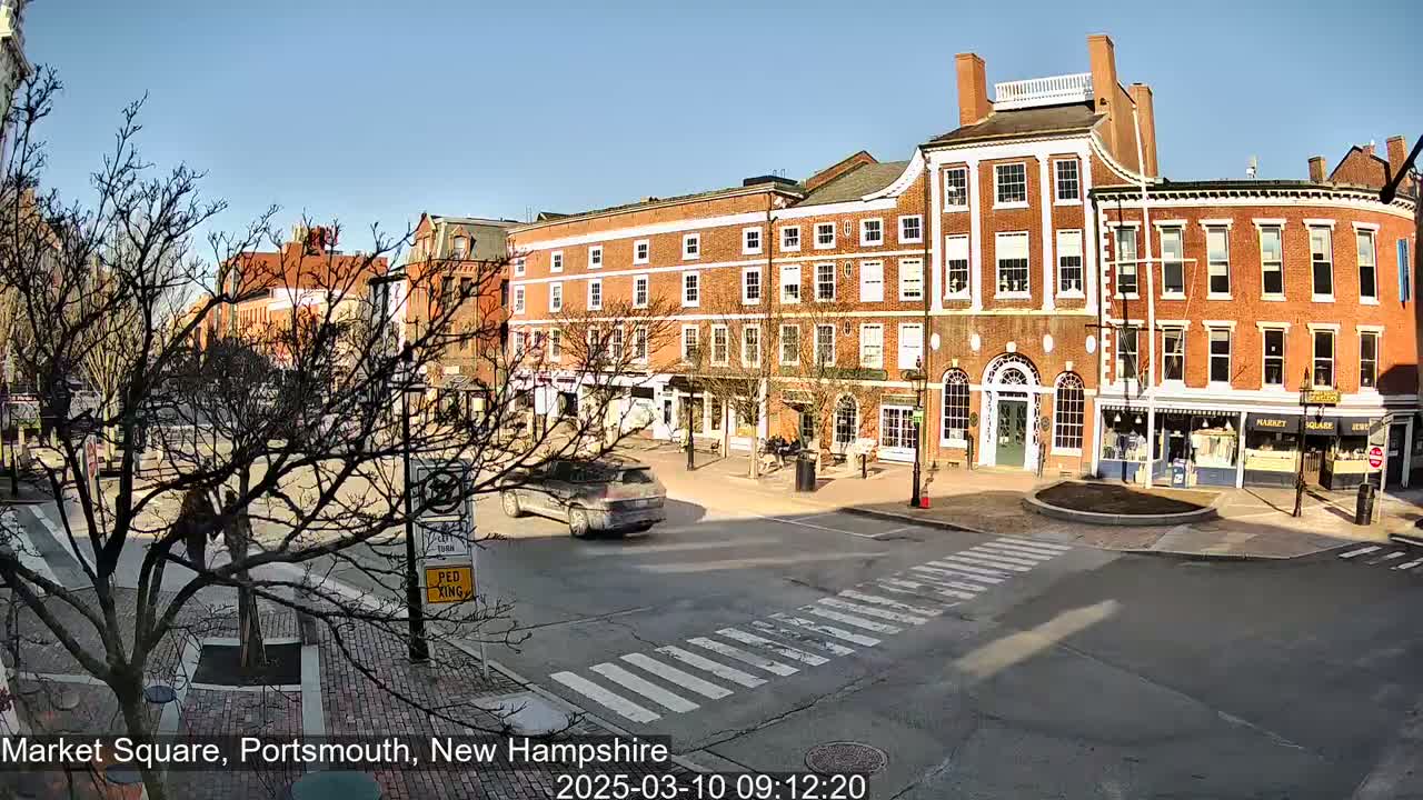 A sunny day in a town square shows several brick buildings, a pedestrian crossing, and a car driving through the square.