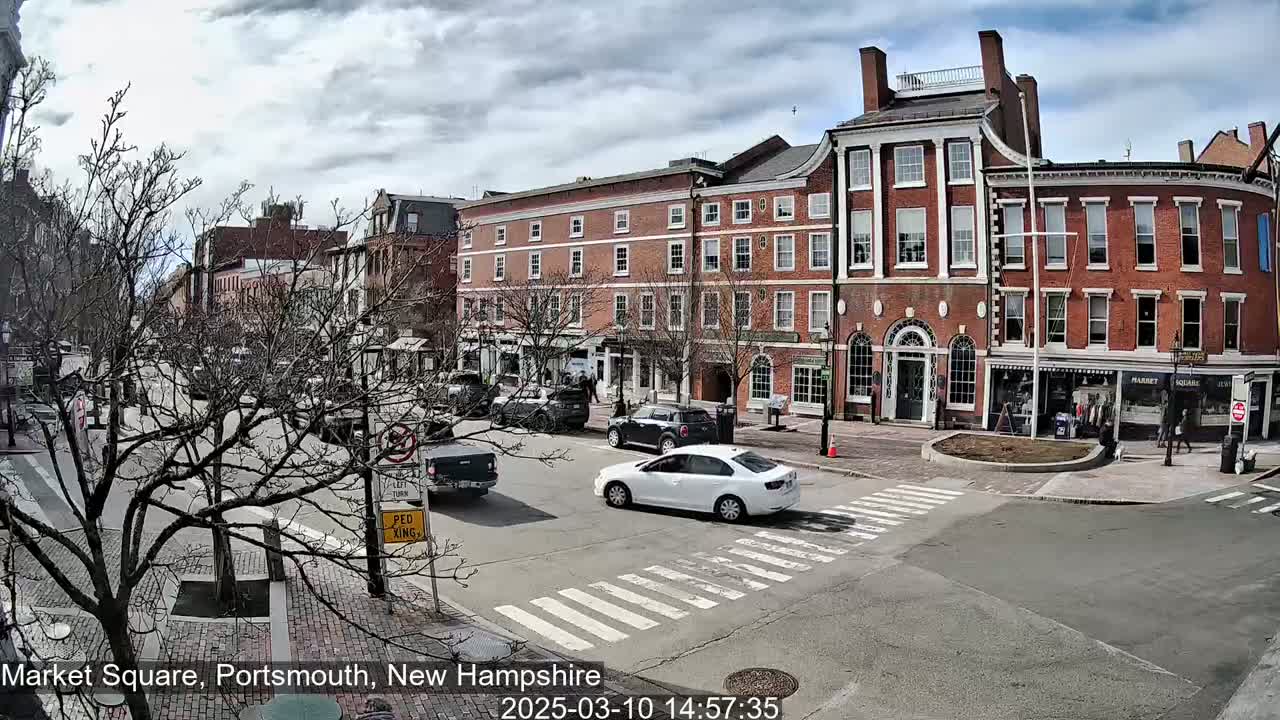 A partly cloudy day in a city square with several brick buildings, bare trees, and a few cars driving through a crosswalk.