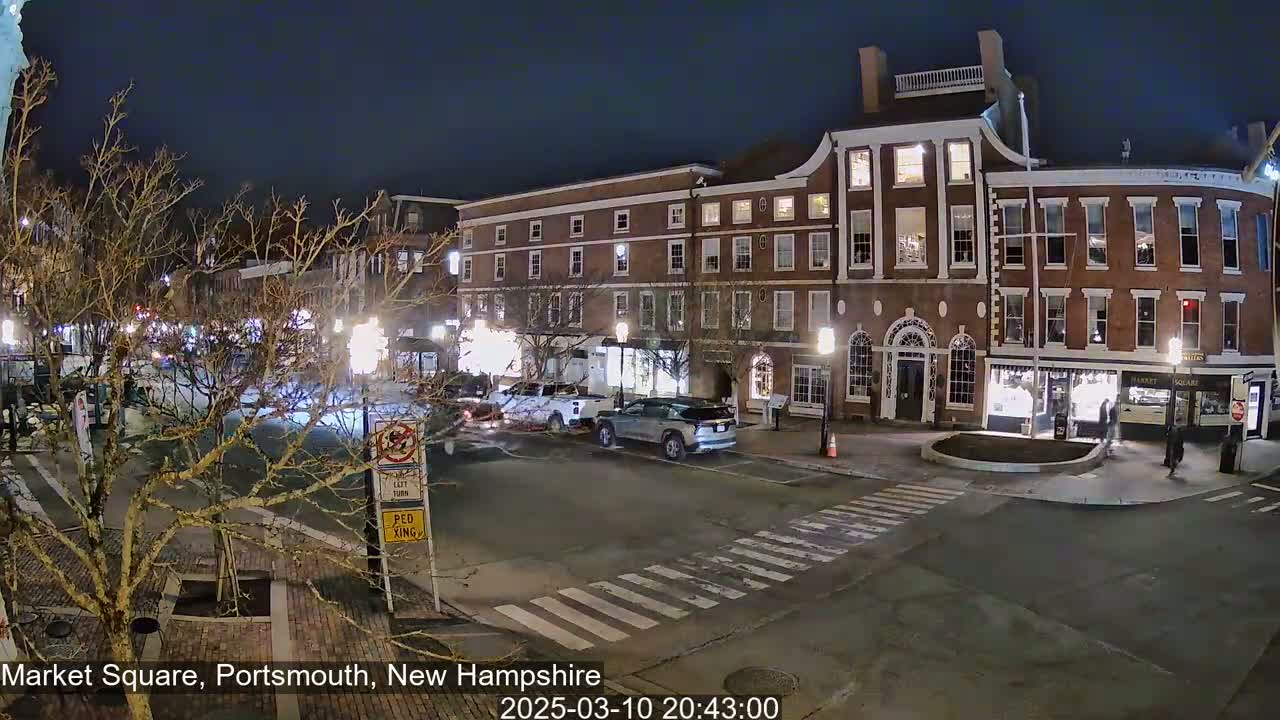 A nighttime street scene shows several brick buildings with illuminated windows, a few cars, and pedestrians on a crosswalk in a city square under a clear, dark sky.