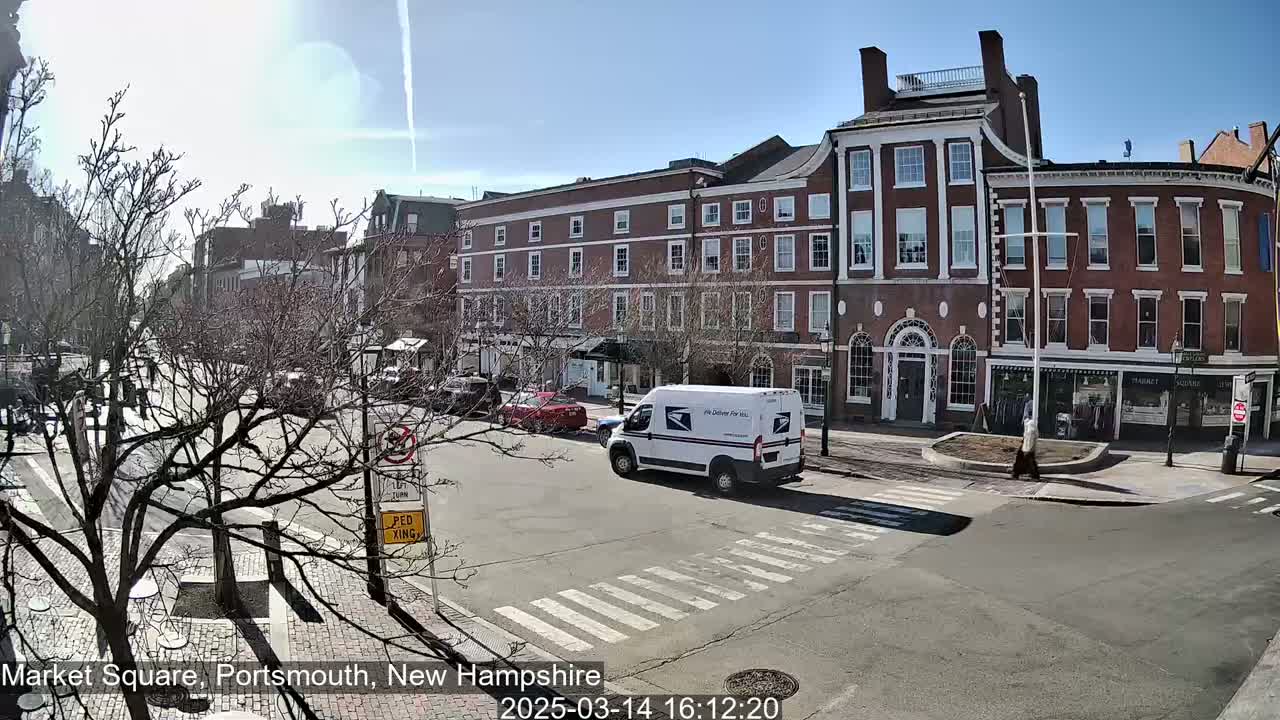 A sunny day in a city square shows a brick building, a white postal truck, and bare trees.