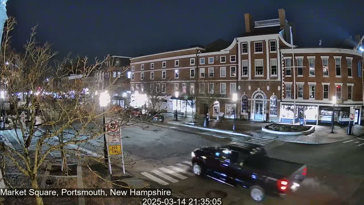 A dark-colored pickup truck drives through a nighttime intersection in front of brick buildings illuminated by streetlights.