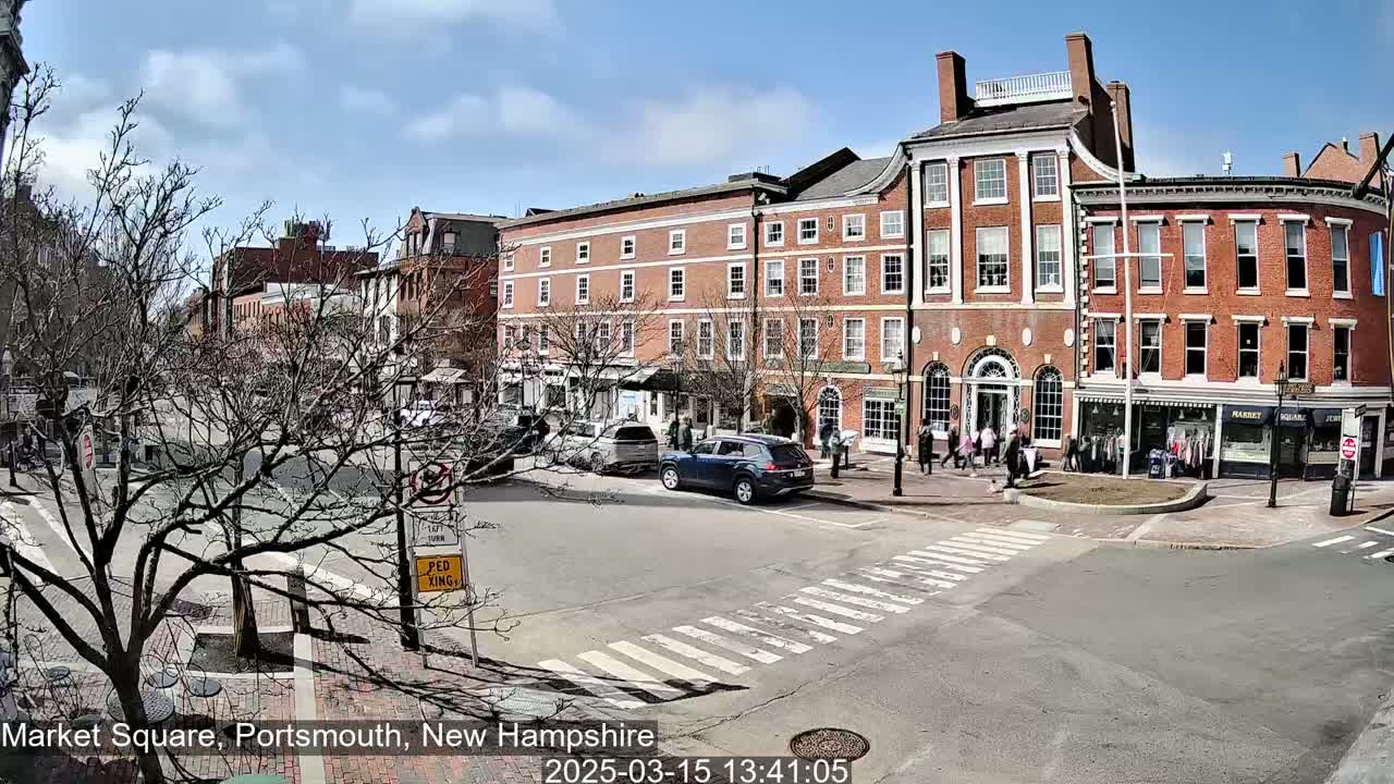A sunny day reveals a town square with several brick buildings, parked cars, and pedestrians walking along a street.