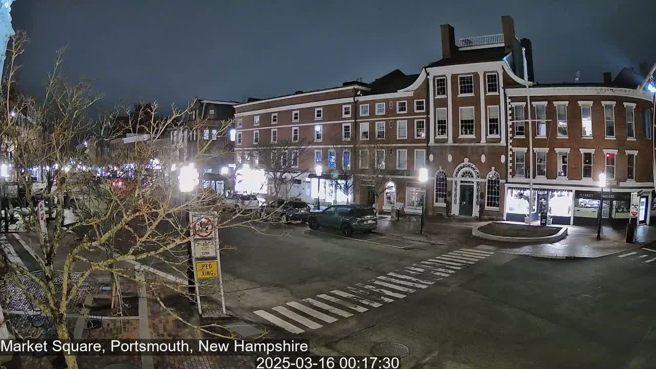 A nighttime view of a brick-lined street with several parked cars, illuminated by streetlights under a dark sky.