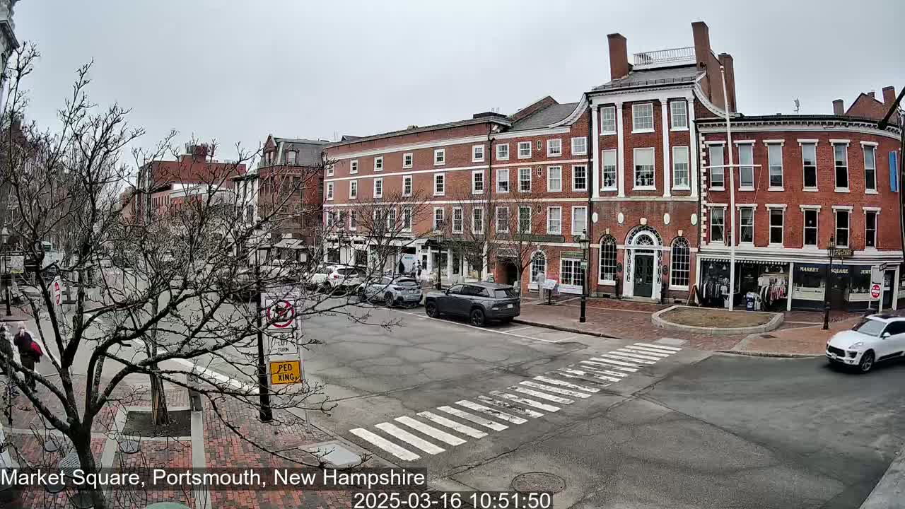 A mostly cloudy day reveals a brick street scene with several multi-story buildings and a few cars parked and driving along the road.