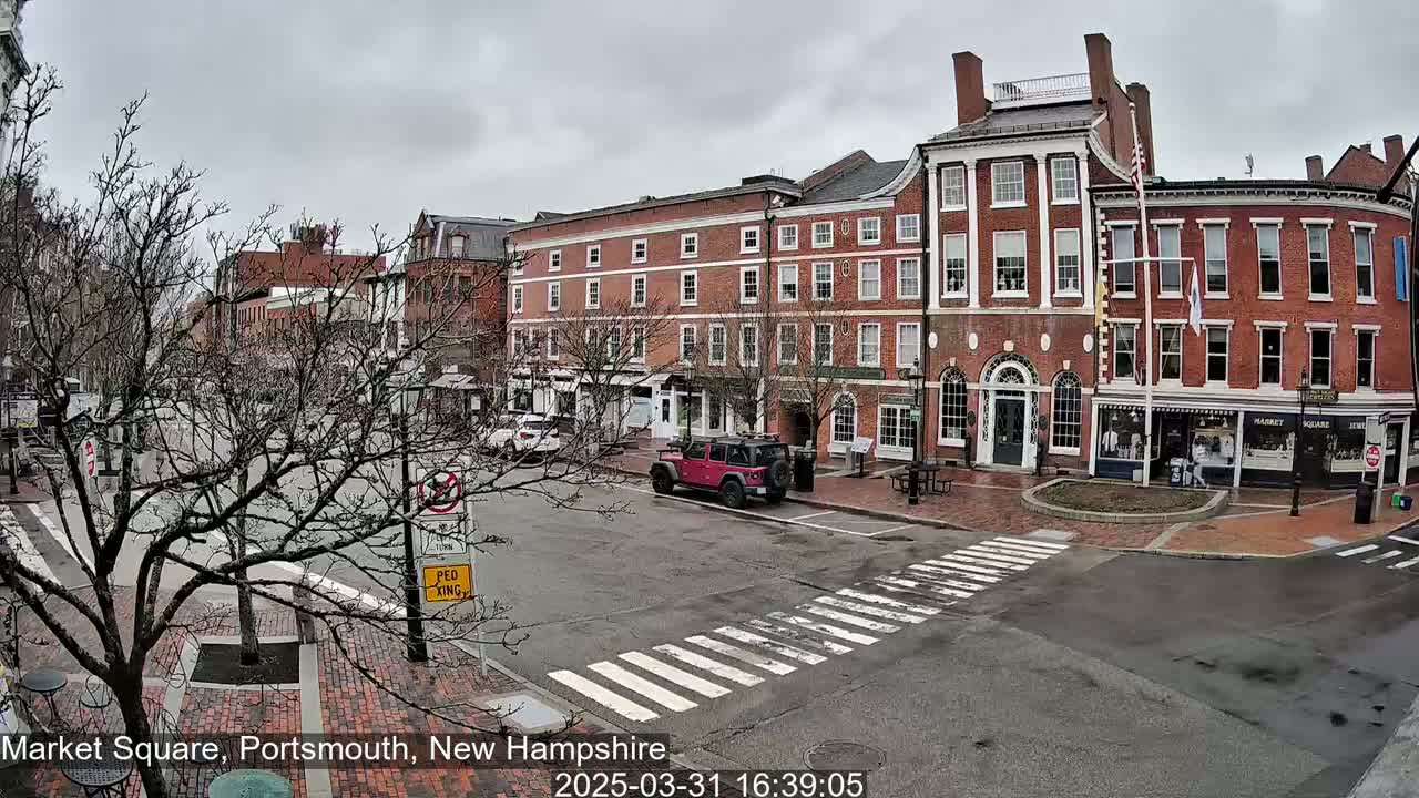 A mostly cloudy day in a brick-lined town square with several cars parked and a red Jeep in the street.