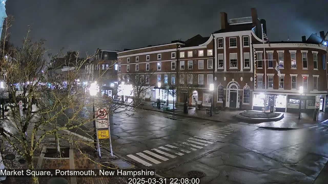 A wet, nighttime street scene shows a row of brick buildings with streetlights illuminating a mostly empty town square.