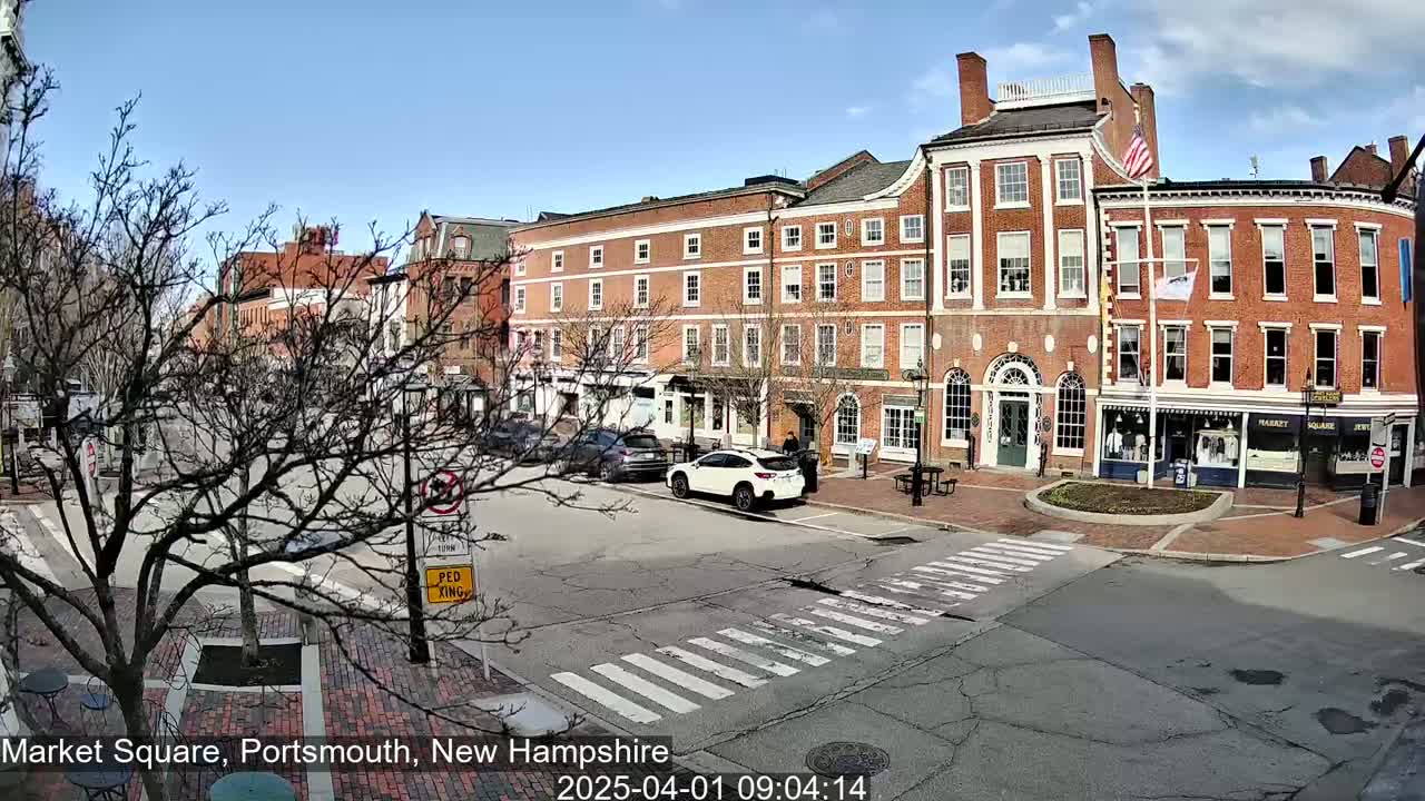 A sunny day in a brick-lined town square, with several cars parked and a few pedestrians visible near multi-story buildings.