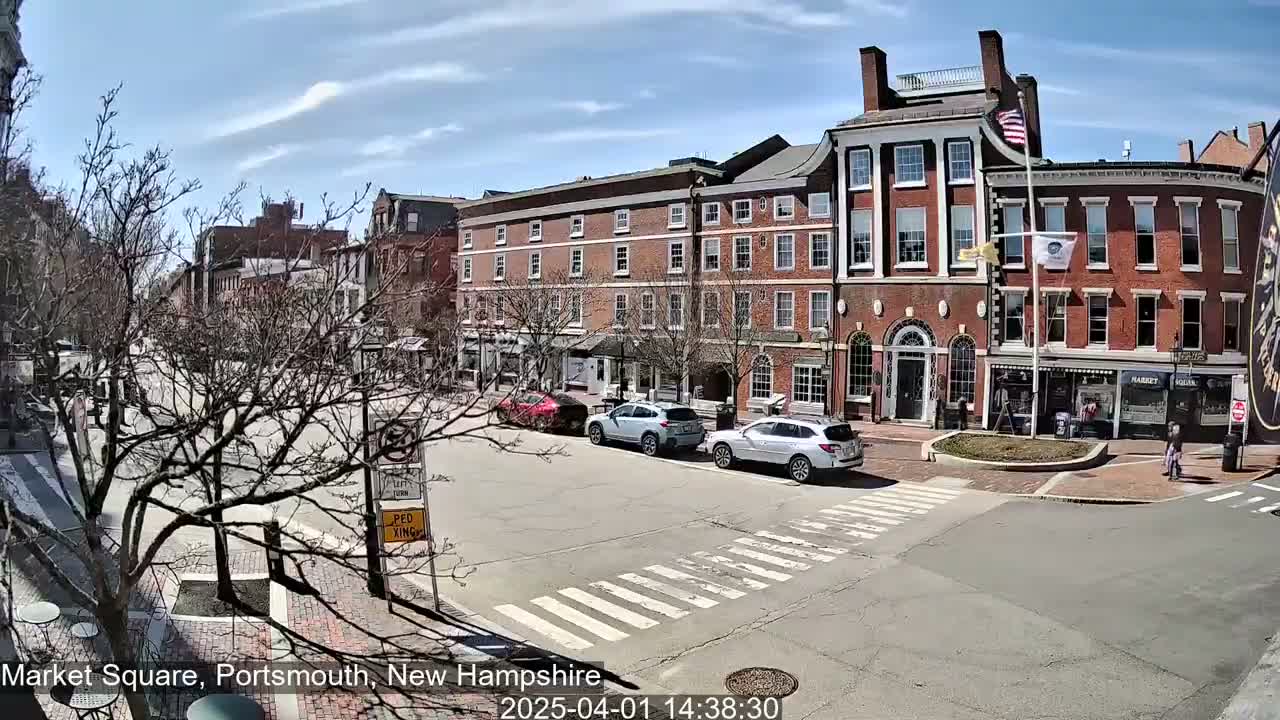 A sunny day reveals a city square with several brick buildings, bare trees, and a few cars parked along the street.