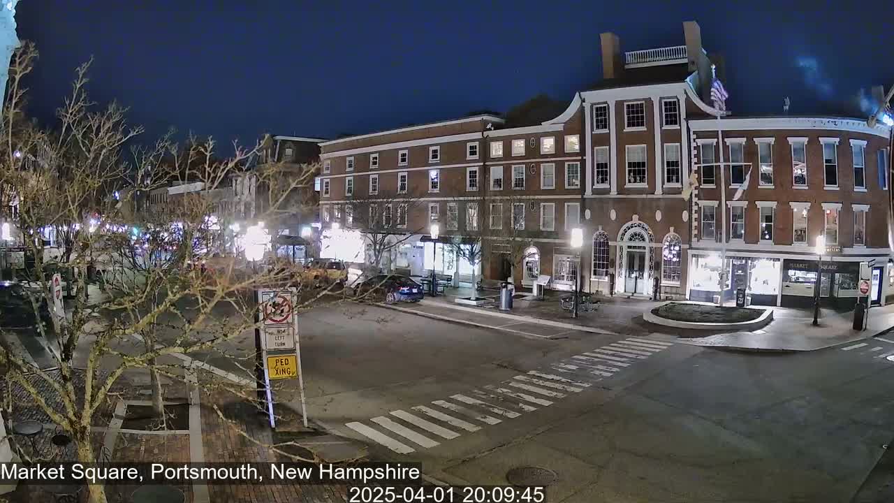 A nighttime view of a town square with brick buildings, streetlights, and a few cars, under a clear, dark sky.