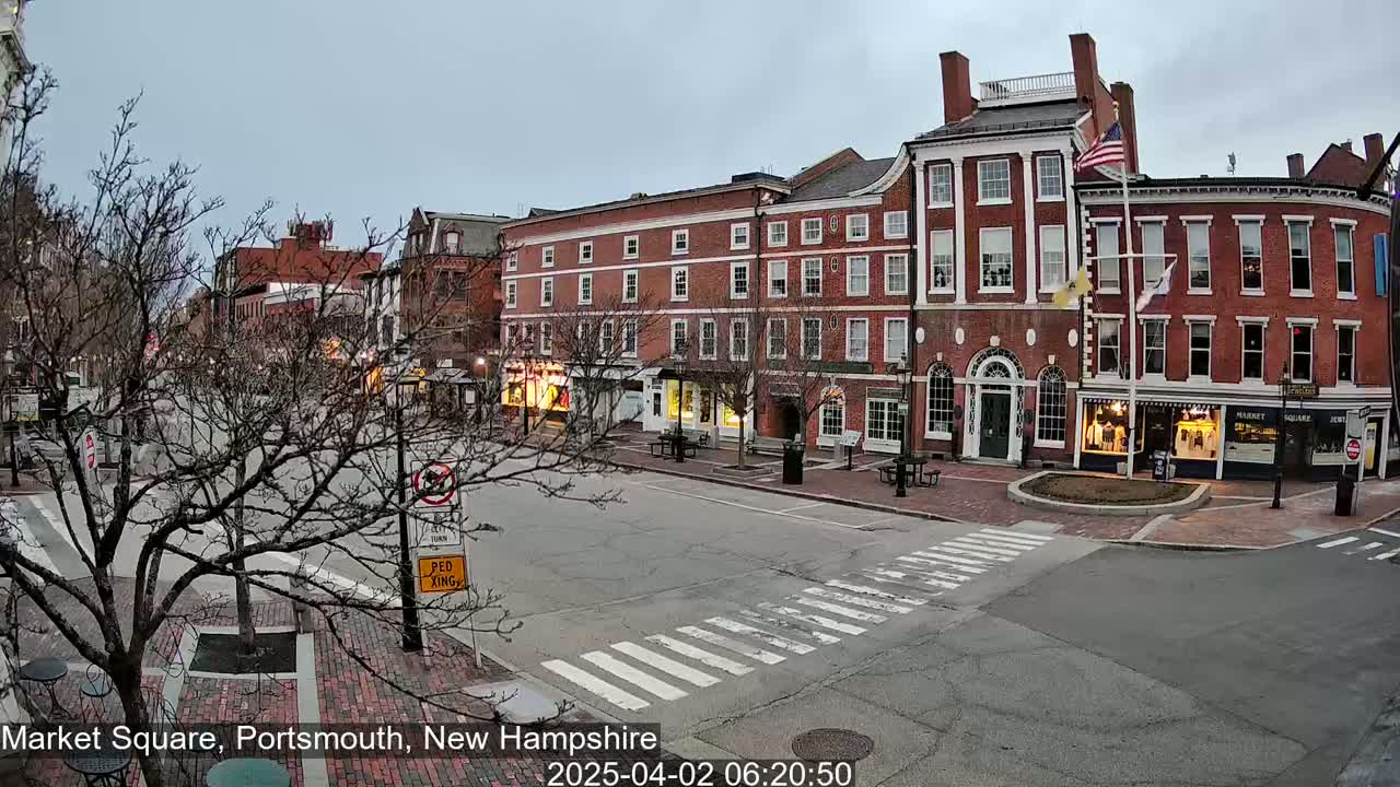 A mostly empty brick-lined town square, under an overcast sky, features several multi-story buildings with various shops at street level.