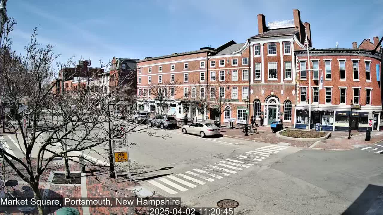 A sunny day in a brick-lined town square with several cars parked and a few pedestrians walking near brick buildings.