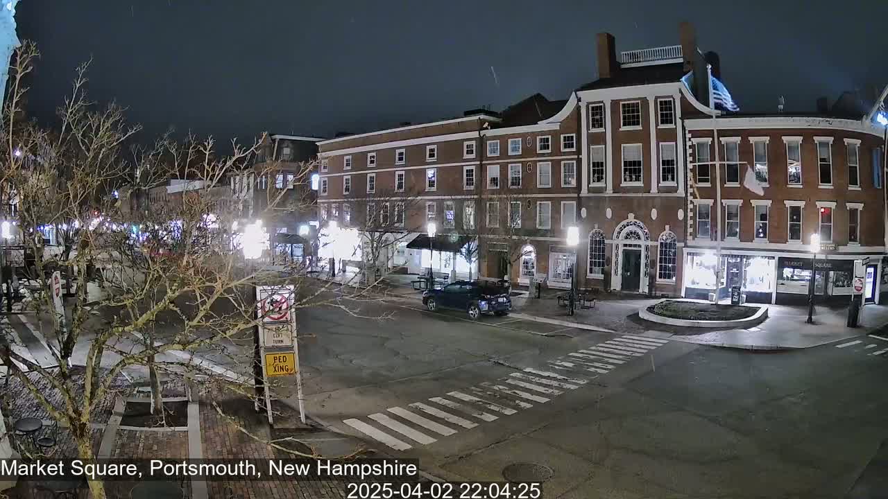 A nighttime view of a snow-dusted town square with brick buildings, streetlights, and a single vehicle.