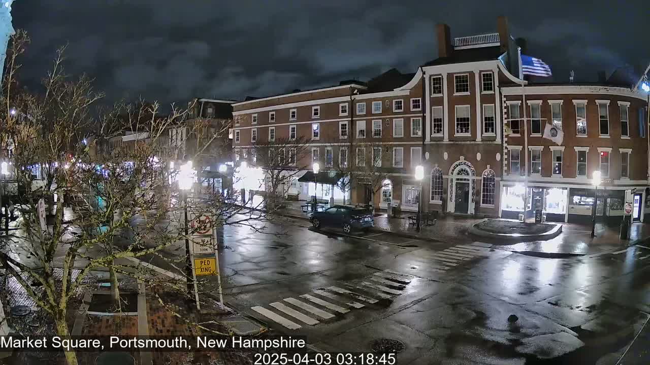 A wet, nighttime street scene shows a brick building with illuminated storefronts and a few vehicles in a town square.