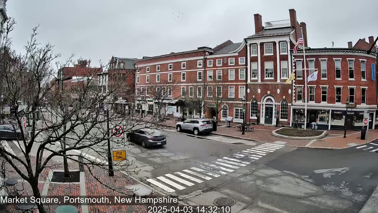 A brick-lined town square with several multi-story buildings, bare trees, and a few cars is visible under an overcast sky.