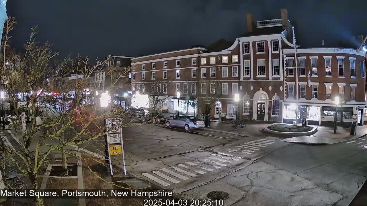 A nighttime view of a brick building in a town square, with a few cars and pedestrians present under a dark sky.
