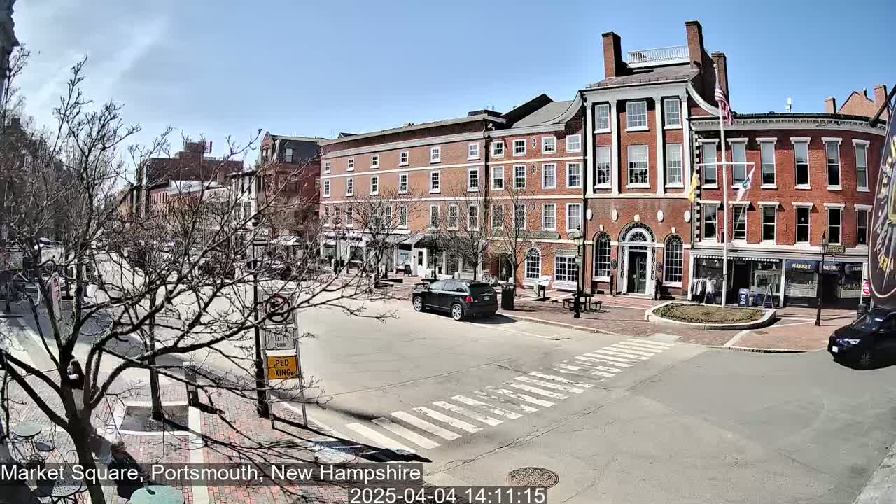 A sunny day in a town square shows several brick buildings, a few cars, and bare trees.