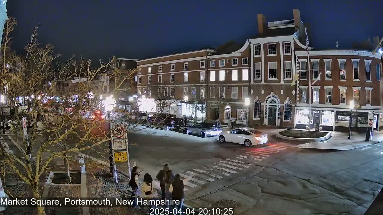 A nighttime street scene shows a brick building with lit windows, cars driving on a street, and pedestrians crossing at a crosswalk under a clear, dark sky.