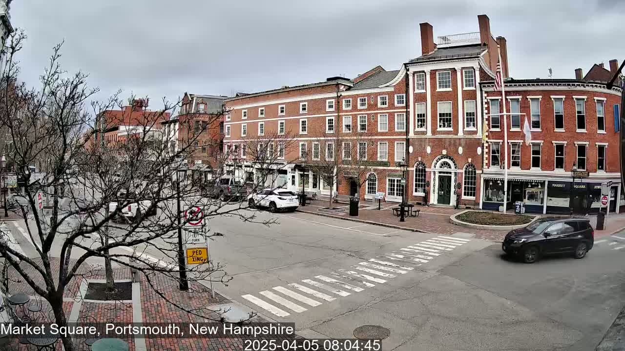 A mostly cloudy day shows a brick-lined city square with several cars and bare trees.