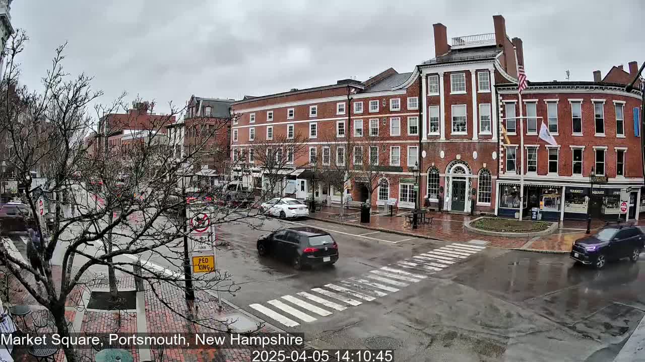 A rainy day scene shows a brick-lined town square with several cars at an intersection in front of historic brick buildings.
