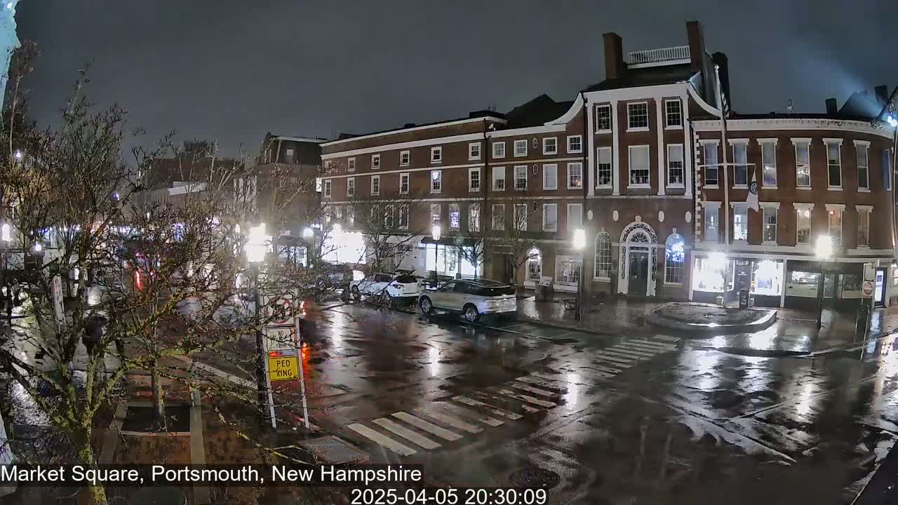 A rain-slicked town square at night, showing several brick buildings with lights on, a few cars parked, and some streetlights illuminating the wet pavement.