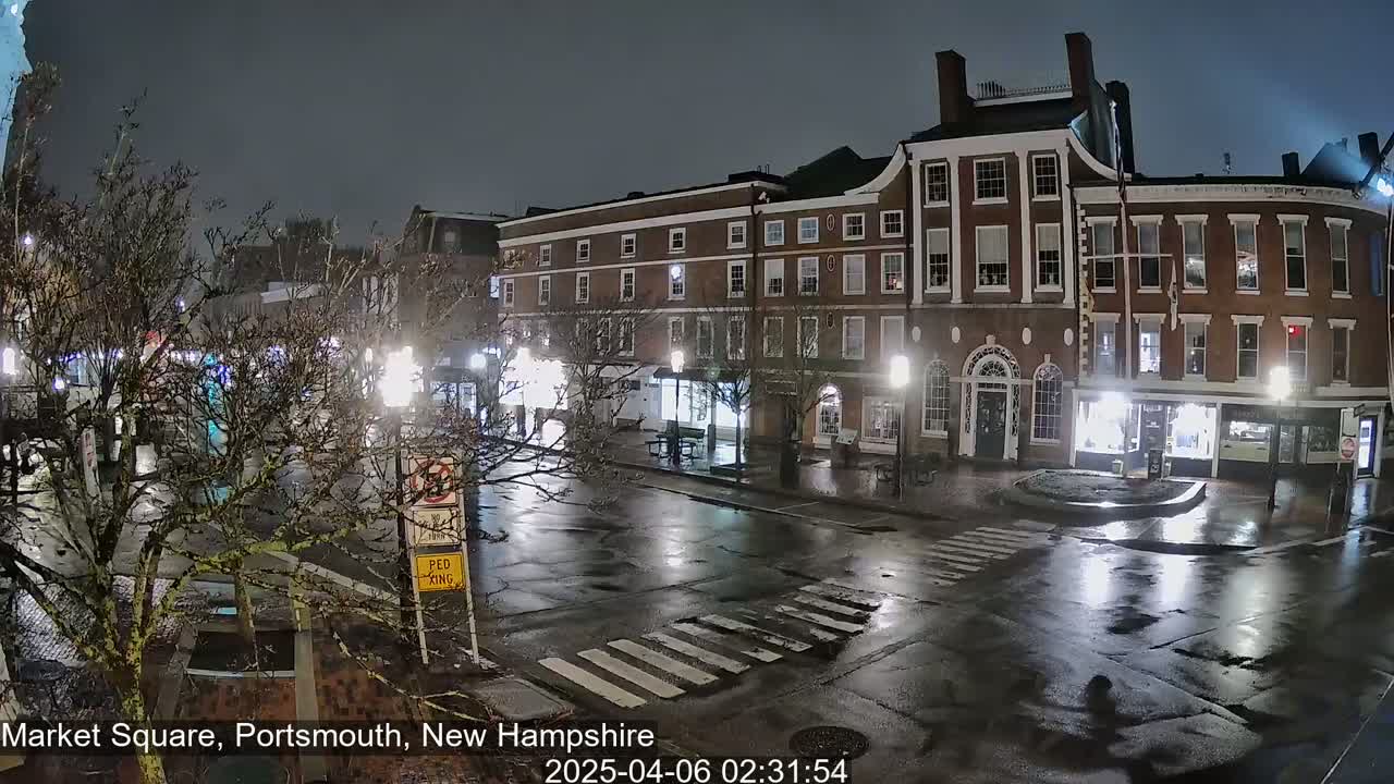 A wet, nighttime street scene shows a brick building with lit windows and streetlights reflecting on the rain-slicked pavement.