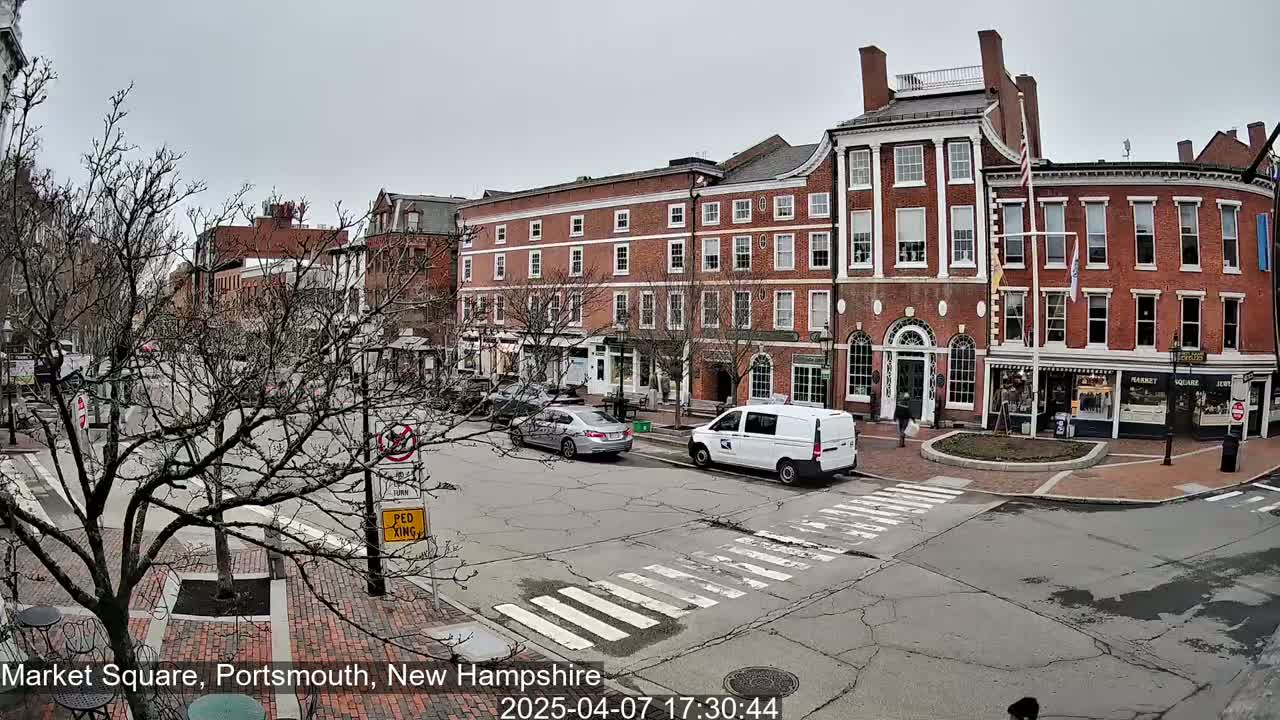 A mostly cloudy day in a town square shows several brick buildings, bare trees, and a few cars and pedestrians on a street with crosswalks.