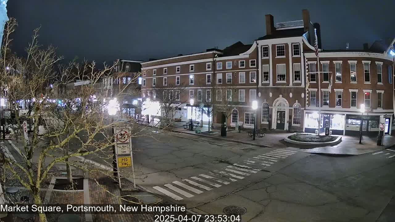 A nighttime view of a mostly empty town square with brick buildings and bare trees under a dark sky.