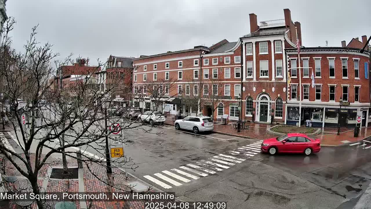 A red car drives through a wet, brick-paved town square lined with multi-story brick buildings under an overcast sky.