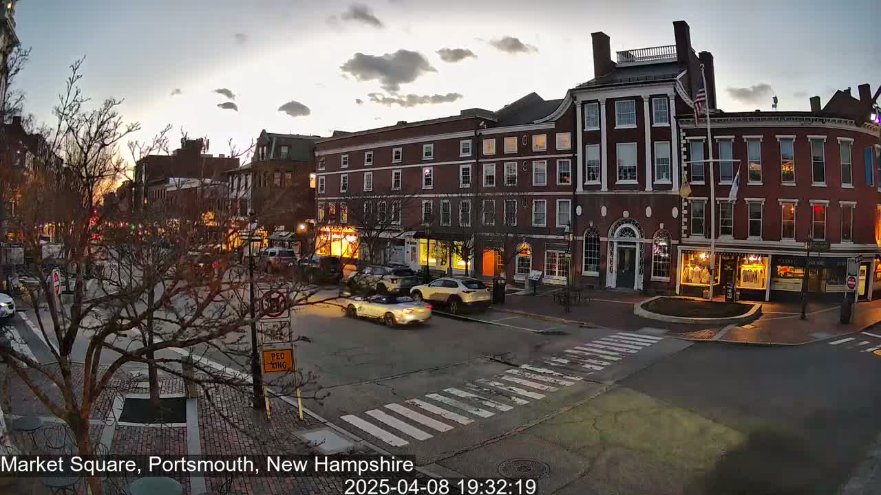 A town square at dusk, with several brick buildings and a few cars on the street under a cloudy sky.