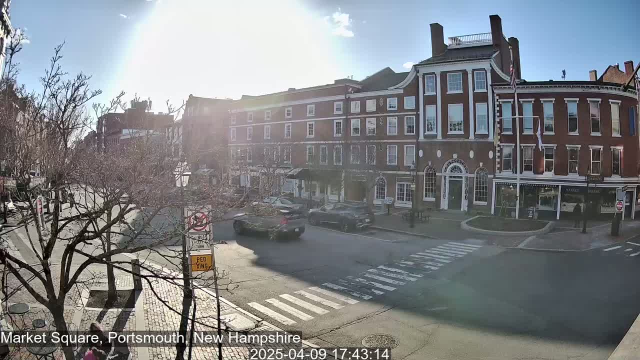 A sunny day reveals a brick street scene with several cars and pedestrians,  bare trees lining the street, and a row of multi-story brick buildings.