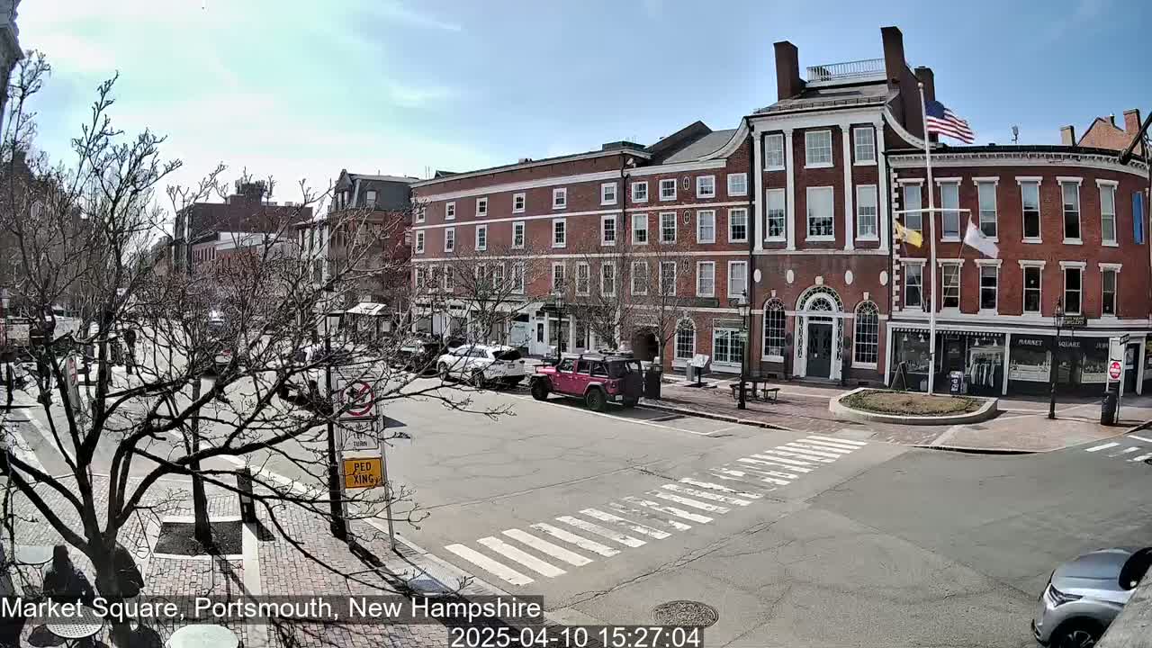 A sunny day reveals a brick-lined town square with several parked cars and sparse pedestrian traffic.
