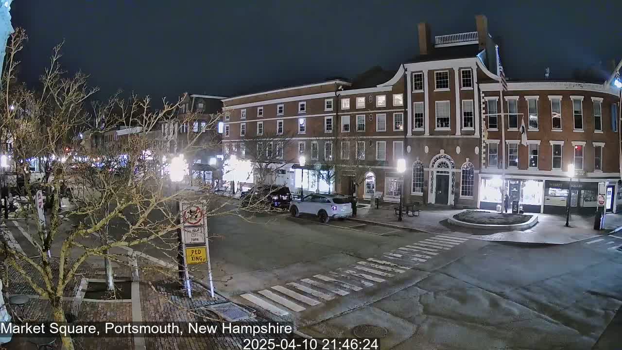 A nighttime view of a brick building-lined street, with a few cars parked and some pedestrians visible under dark, clear skies.