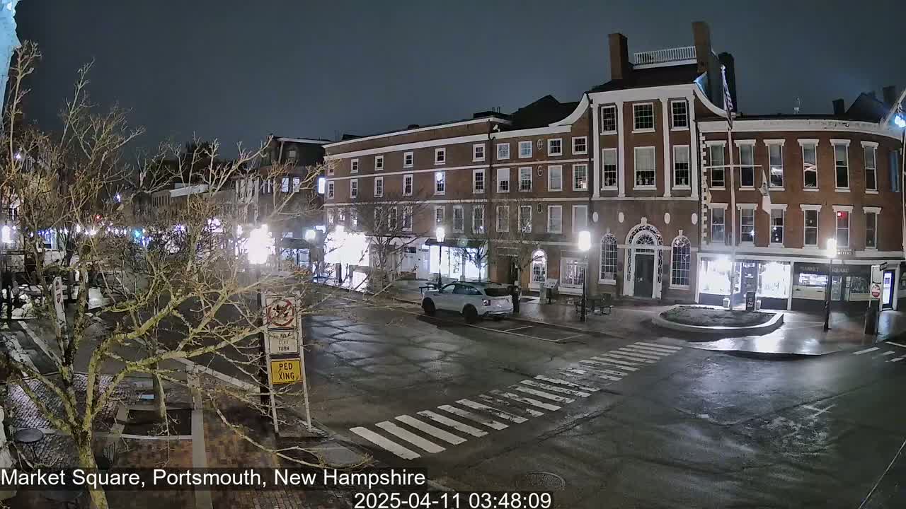 A nighttime view of a town square with several brick buildings, a few cars, and bare trees under a dark sky.
