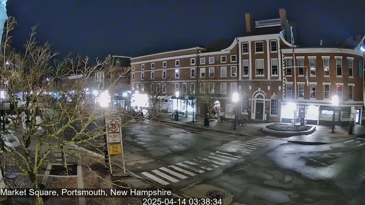 A nighttime view of a town square with brick buildings, streetlights, and a wet street.
