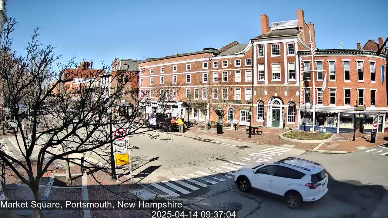 A sunny day reveals a brick-lined town square with several multi-story brick buildings, bare trees, and a white SUV driving through a crosswalk.