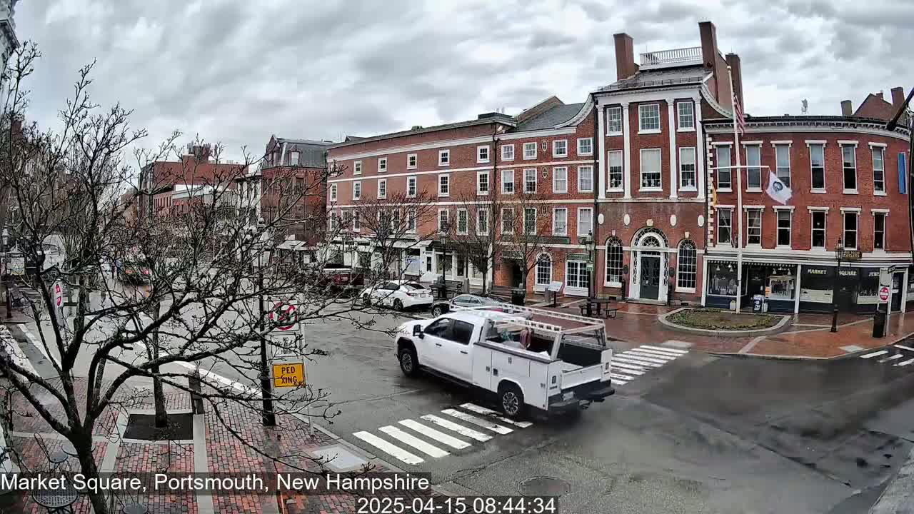 A wet brick town square with several multi-story brick buildings under a cloudy sky, and a white pickup truck driving through a crosswalk.