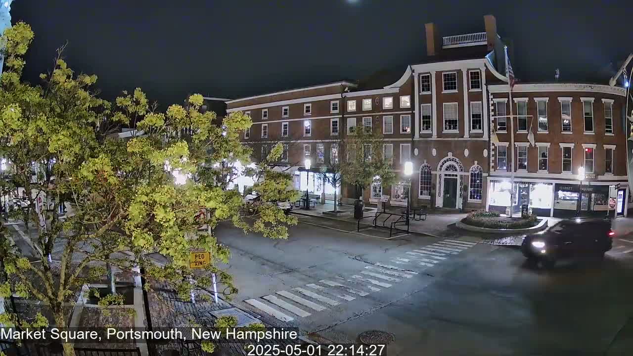 A dark-colored SUV drives through a pedestrian crossing in a town square at night, passing brick buildings illuminated by streetlights under a clear sky.