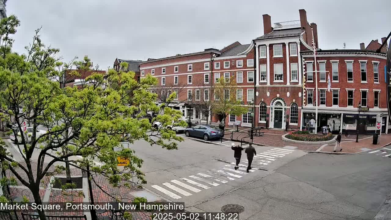 Two people cross a street in front of brick buildings on an overcast day.
